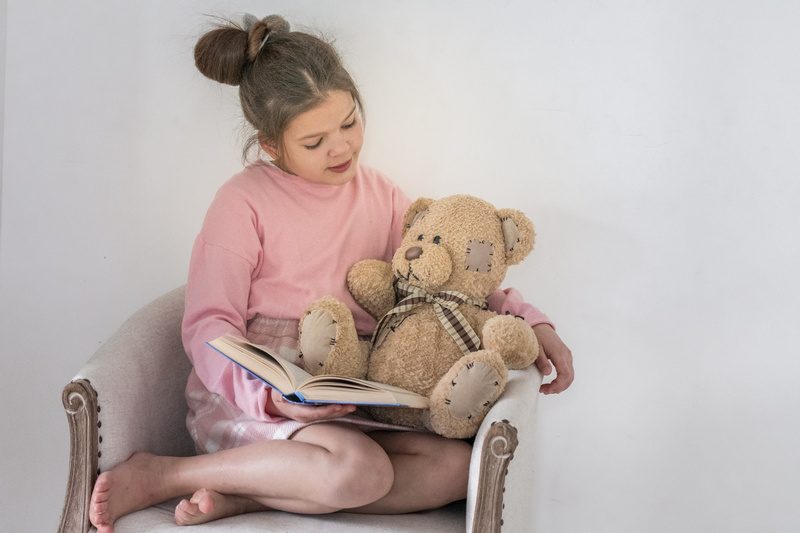 Dziewczynka siedząca w fotelu czytająca książkę pluszowemu misiowi